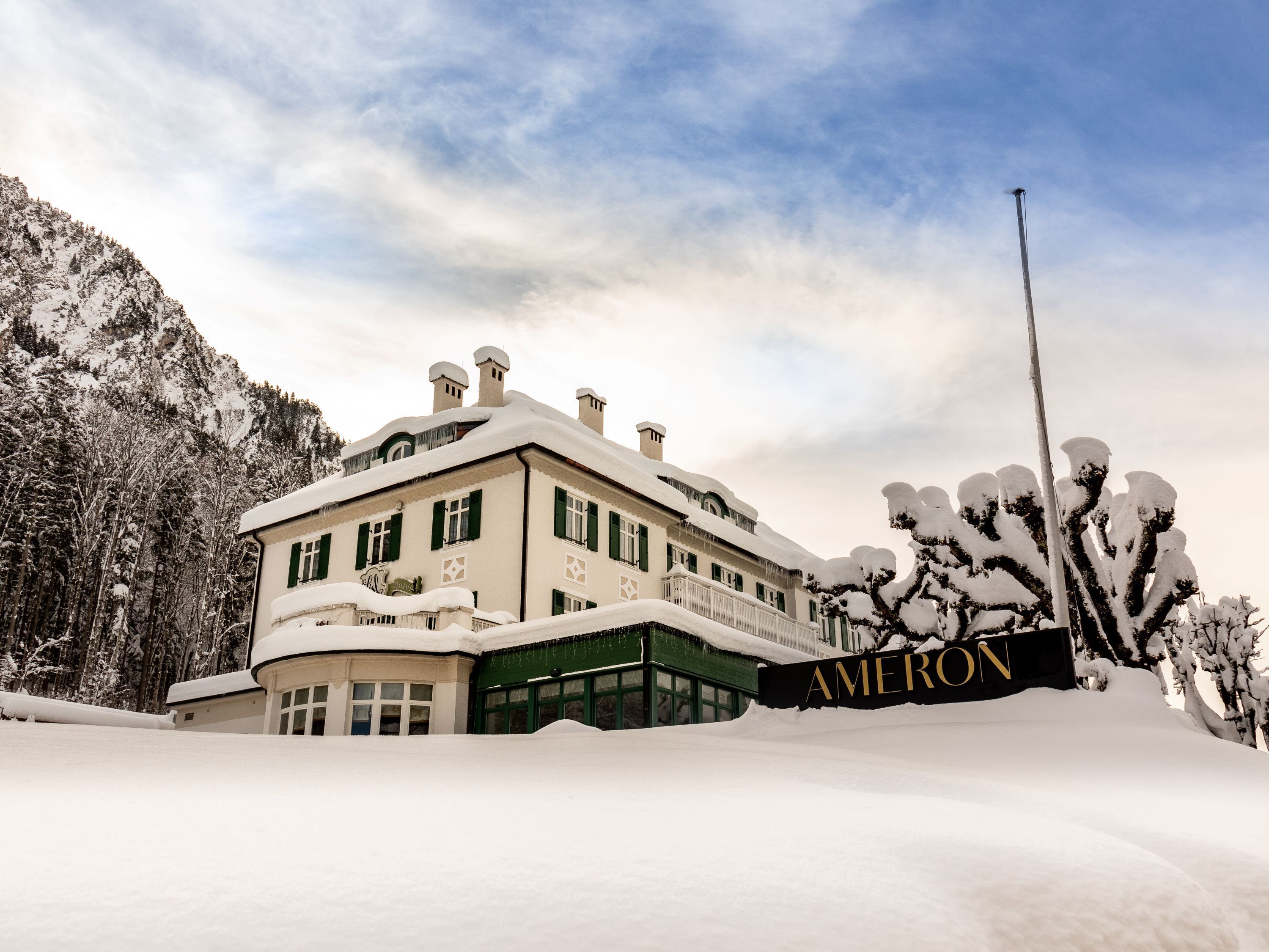 A large, snow-covered building with a prominent "AMERON" sign, surrounded by snow-laden trees and mountains.