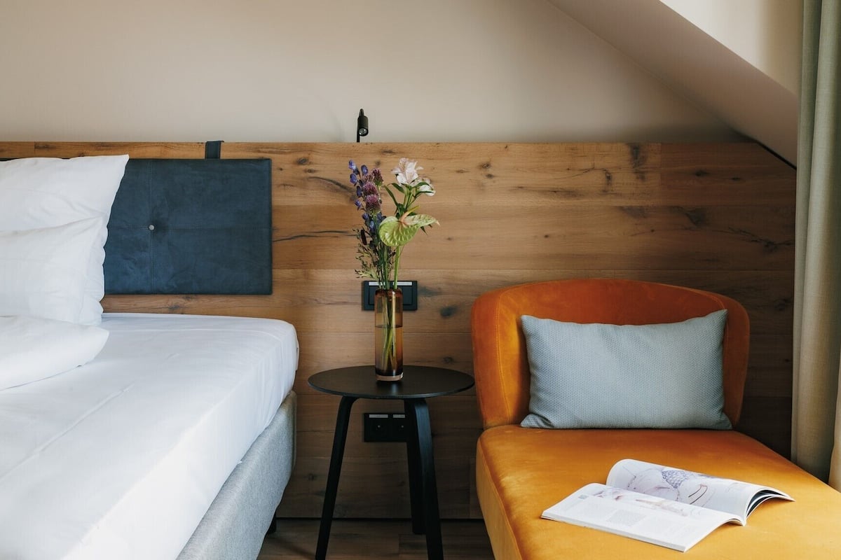 A hotel room with a bed, a small table with a vase of flowers, a chair with a cushion, and a magazine on the table.