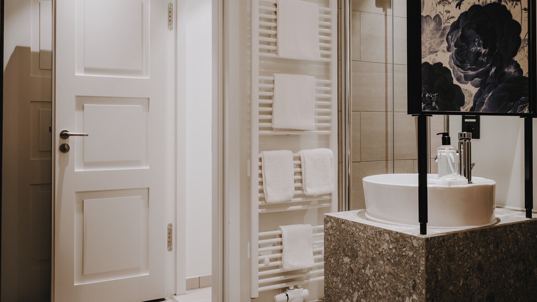 Un bagno moderno con una porta bianca, un appendi asciugamani pieno di asciugamani, un lavandino con piano in marmo e uno specchio con decorazioni floreali.