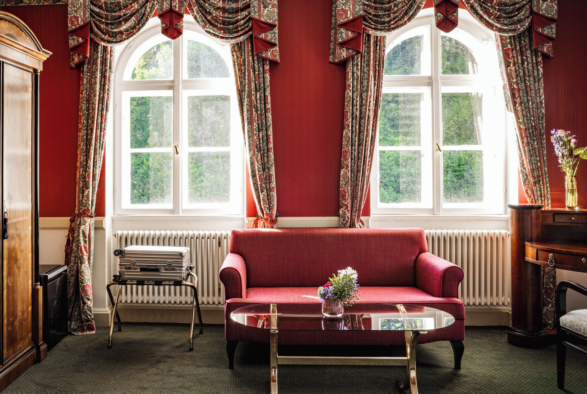 Una habitación con sofá rojo, una mesita de centro de cristal, un radiador y cortinas.