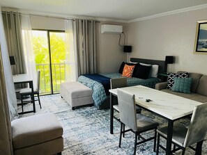 A hotel room with a bed, a desk, a dining table, and a sofa. There is a window with curtains, an air conditioning unit, and a painting on the wall. - Coral Beach Hotel and Condominiums (Freeport)