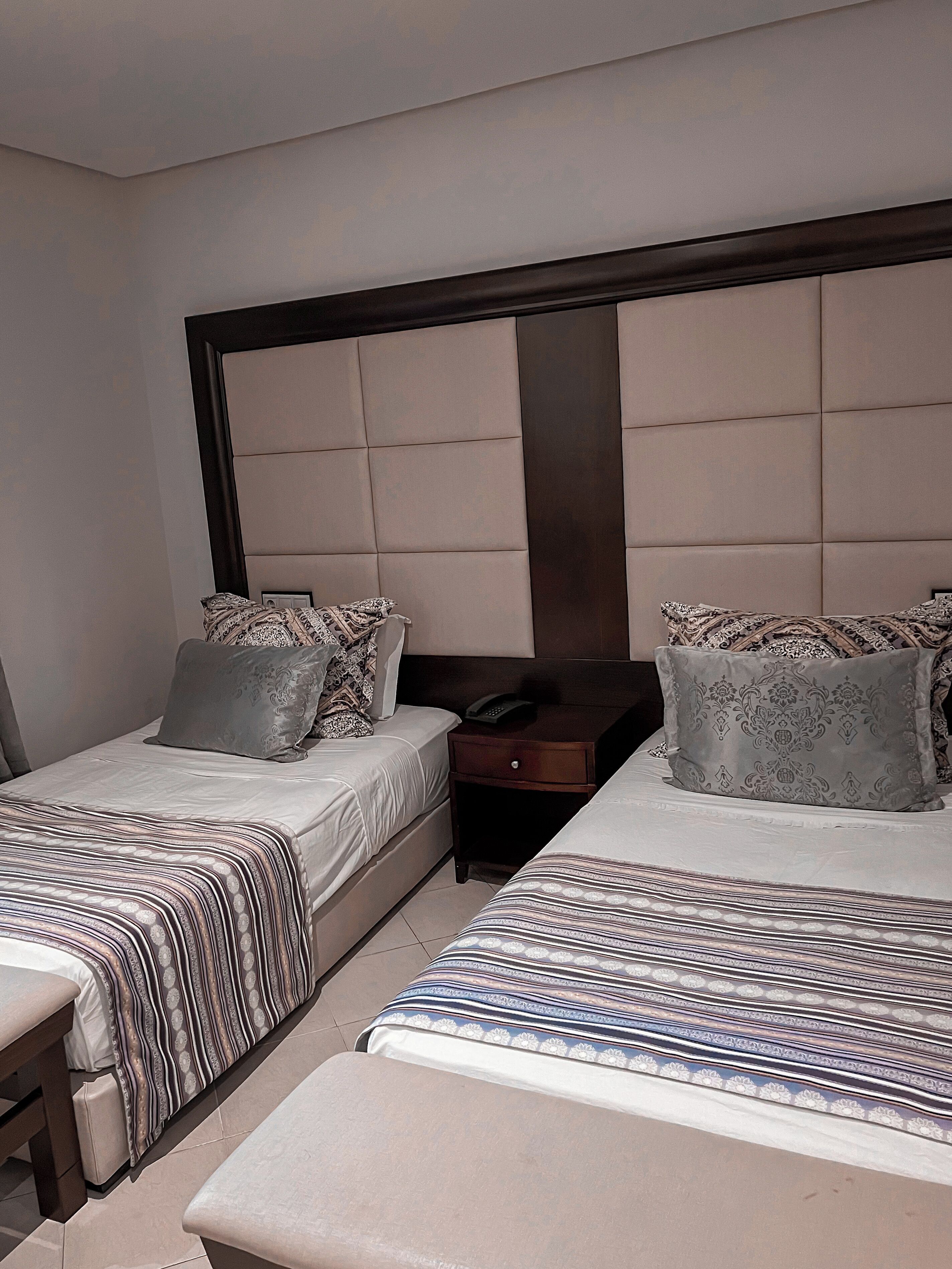 A hotel room with two beds, a nightstand, and a bench.