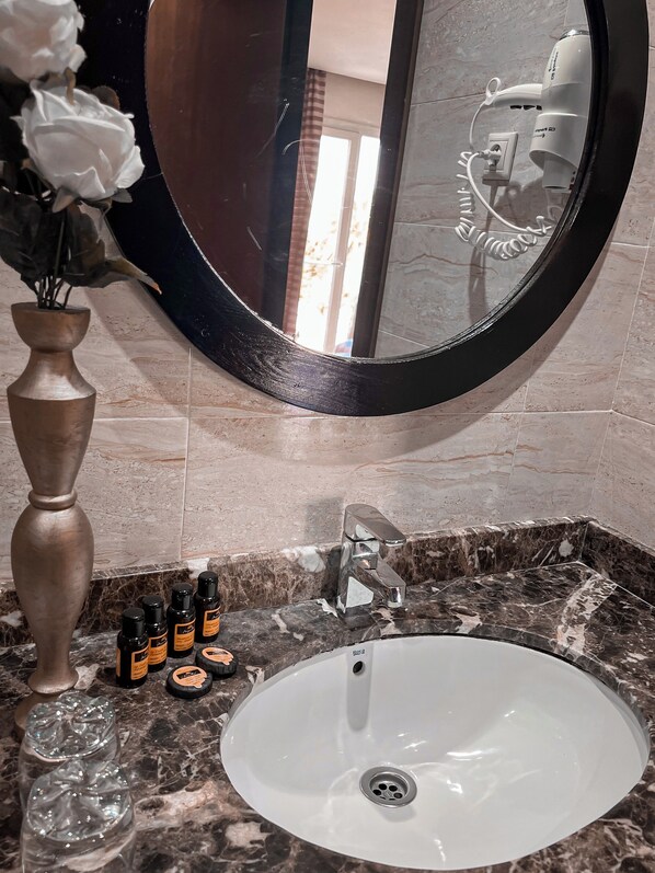 A bathroom with a white sink, a round mirror, a vase with flowers, and a marble countertop.