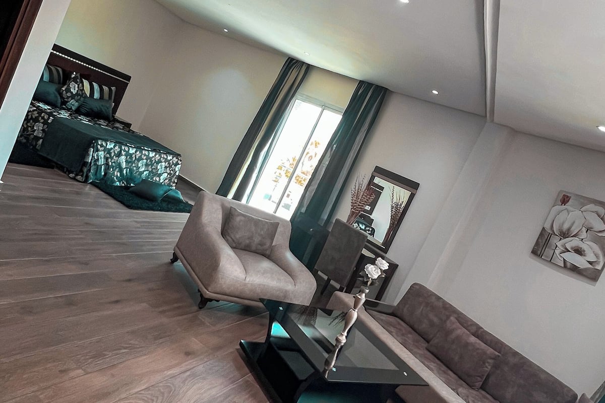 Ein modernes Wohnzimmer mit einer Couch, einem Sessel und einem Glastisch. Im Hintergrund ist ein Bett mit grüner Bettwäsche zu sehen.