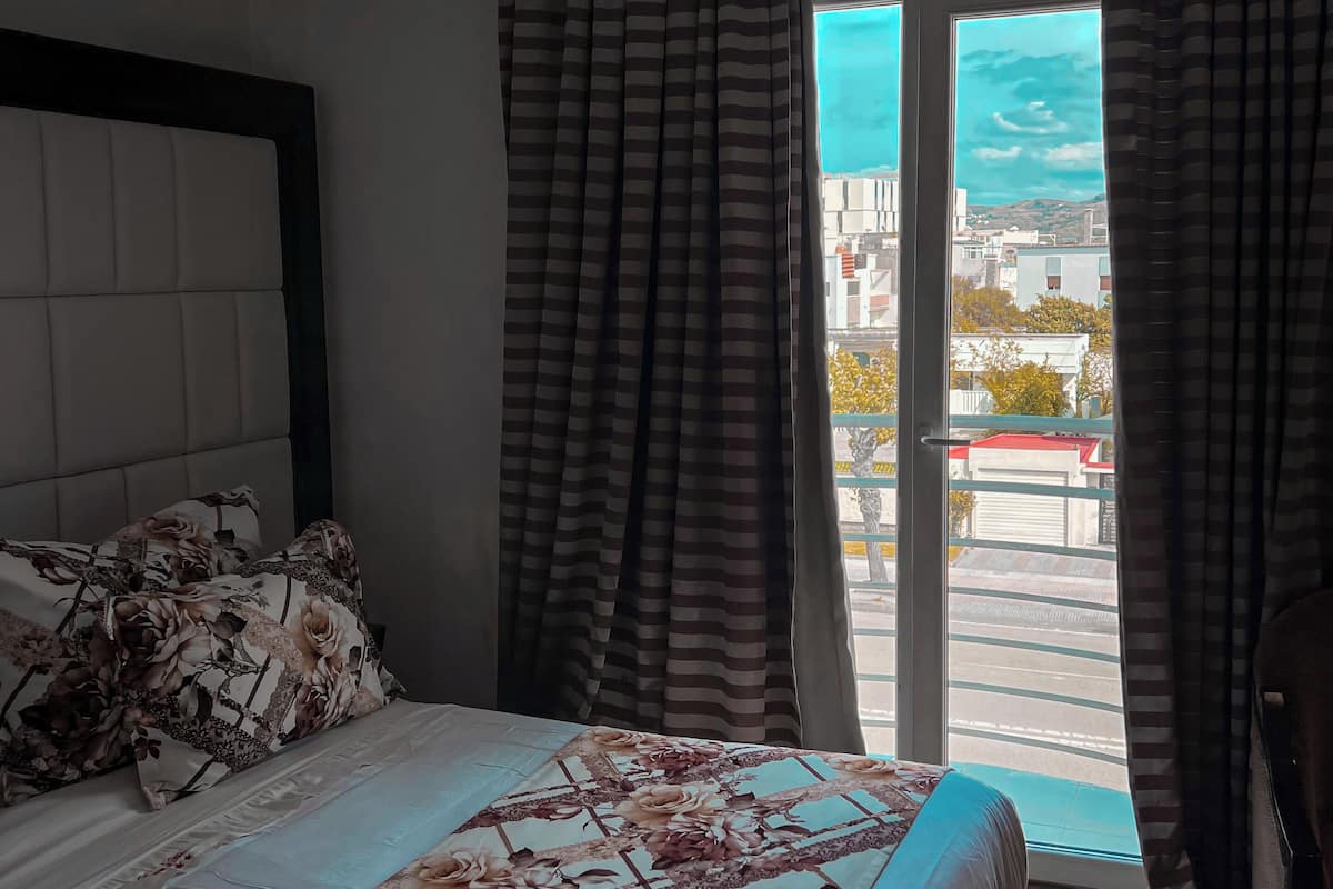 Ein Schlafzimmer mit Bett, Kopfteil, Vorhängen und Blick auf die Stadt durch ein Fenster.
