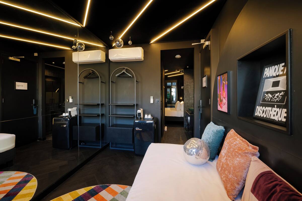 A modern hotel room with a large bed, a mirror, a wardrobe, a small desk, and a bathroom with a shower and toilet.