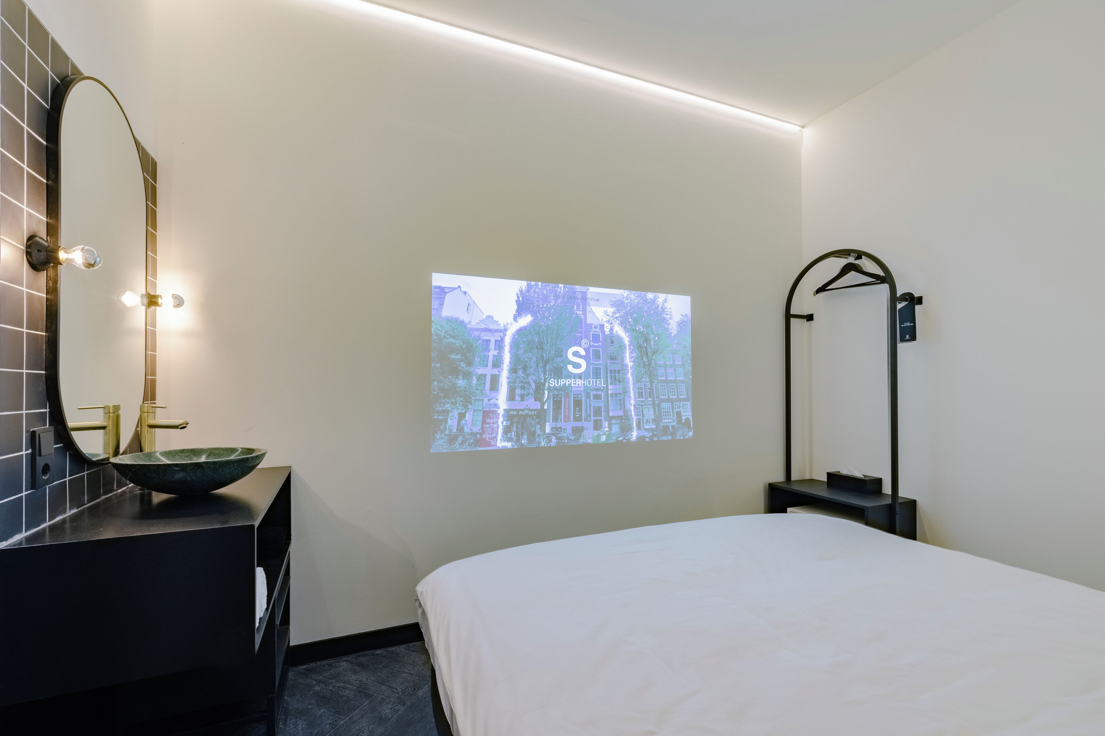 Une chambre d’hôtel avec un lit, une salle de bain équipée d’un lavabo et d’un miroir, et un écran de projection fixé au mur.