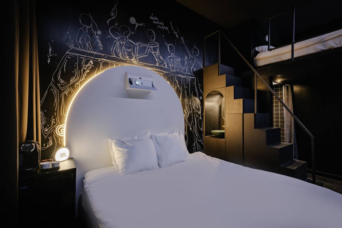 A modern hotel room with a large bed, a headboard with a lighted design, a staircase, and a wall with a mural.