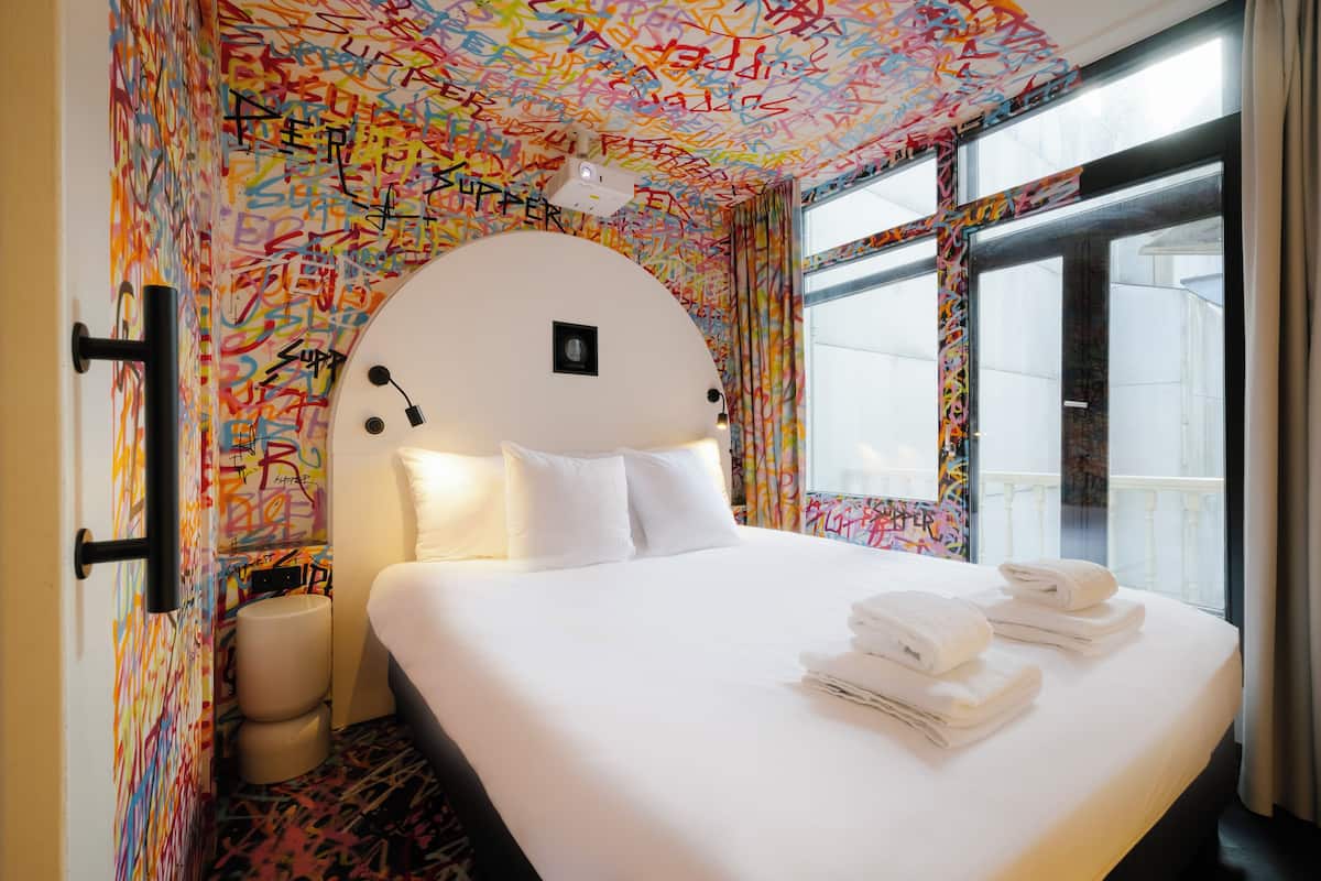 A hotel room with a bed, white linens, a bathroom with a toilet and sink, and a window with a view.