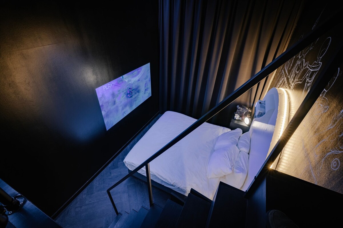 Habitación de hotel moderna con cama, una mesita de noche pequeña y una pantalla mural que muestra una imagen panorámica.