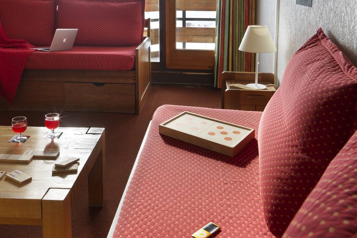 Ein gemütliches Zimmer mit einem roten Sofa, einem Holztisch mit einem Laptop, Gläsern und einem Tablettspiel. Ein Fenster bietet Blick auf eine verschneite Landschaft und eine Seilbahn.