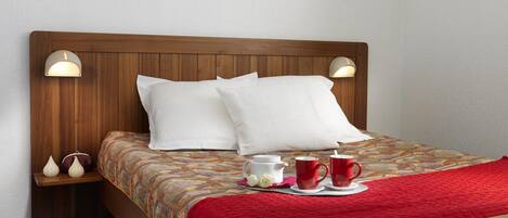 Una cama bien hecha con cabecera de madera, almohadas blancas y una manta roja. Encima de la cama hay un cuadro enmarcado, y una mesita de noche con una tetera y tazas al lado.