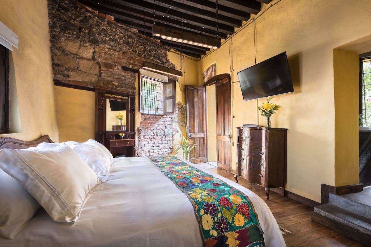 A bedroom with a bed, a wooden dresser, a television, and a brick wall.