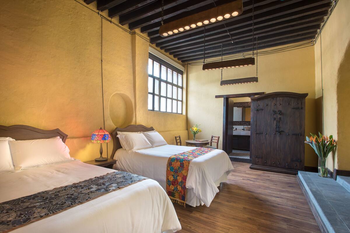 A hotel room with two beds, wooden flooring, a large window, a wooden wardrobe, and a small table with a vase of flowers.