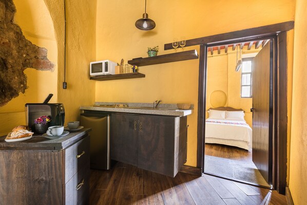 A compact kitchen with a microwave, coffee maker, and a small dining area. A bed is visible through an open door in the adjacent room.