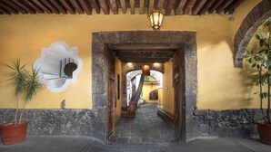Un arco de piedra que da acceso a un pasillo con pared amarilla, plantas en macetas y un farol.