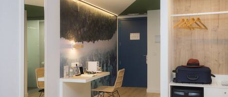 A compact hotel room with a bed, a desk, a chair, a small kitchenette, and a wardrobe with hangers.
