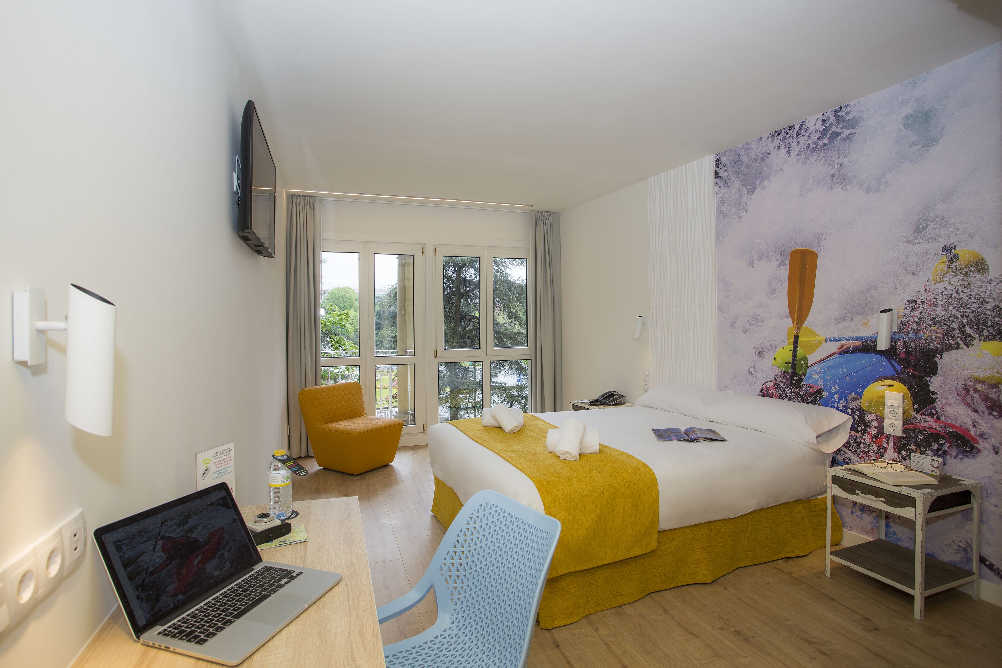 A hotel room with a bed, a desk with a laptop, a chair, a nightstand, and a large window.