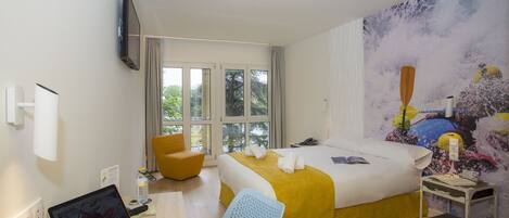 A hotel room with a bed, a desk with a laptop, a chair, a nightstand, and a large window.