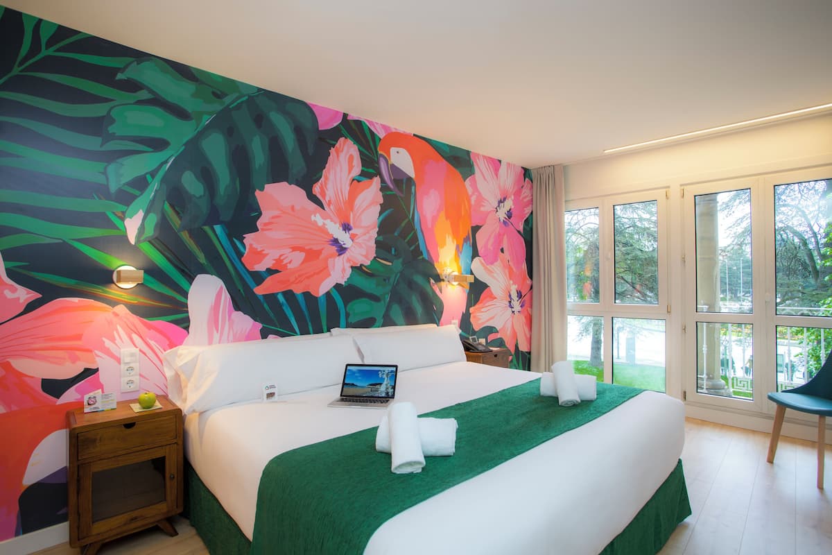 Une chambre d’hôtel avec un grand décor mural floral, un lit recouvert d’une couverture verte, un ordinateur portable posé sur le lit et une chaise près de la fenêtre.