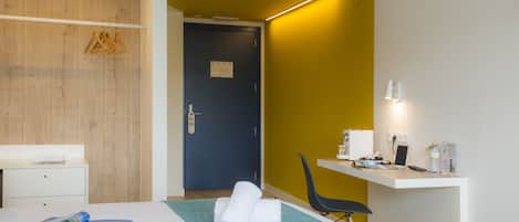 Una habitación de hotel con cama, escritorio, silla, cesto de basura y puerta.