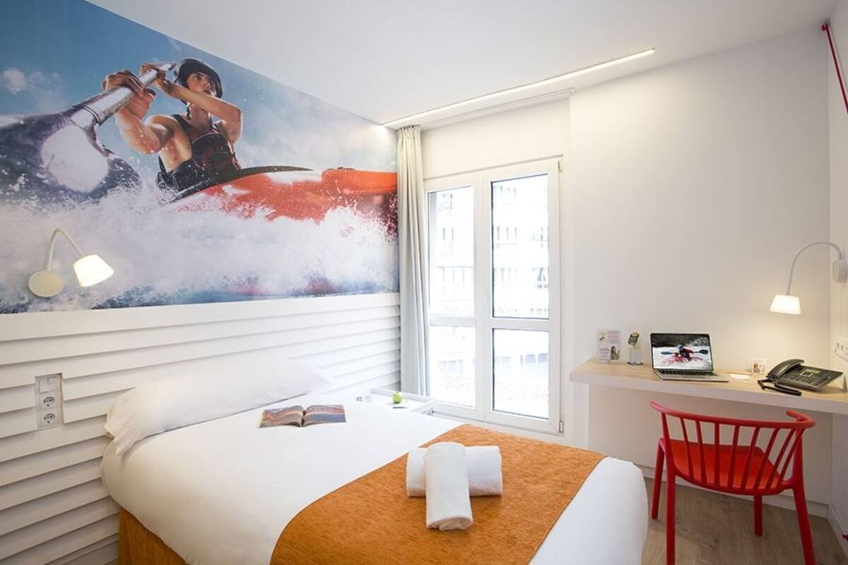 Une chambre d’hôtel avec un lit, un bureau, une chaise rouge, un ordinateur portable et une grande fenêtre.