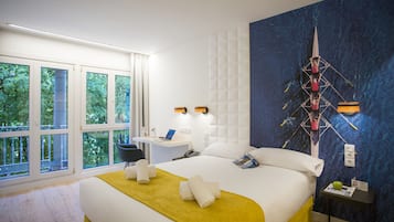 Quarto de hotel com uma cama grande, uma escrivaninha com computador e vista para árvores pelas janelas.