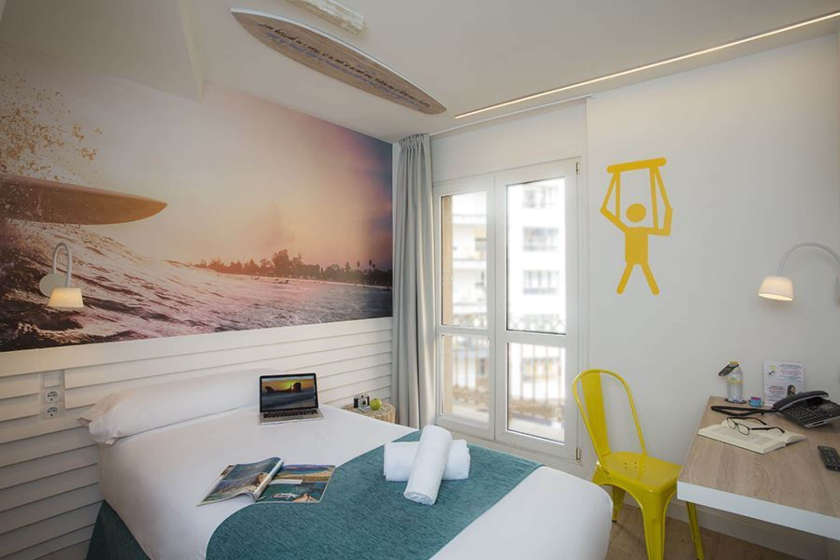 A hotel room with a bed, a desk, a chair, a window, and a mural of a surfer on a wave.