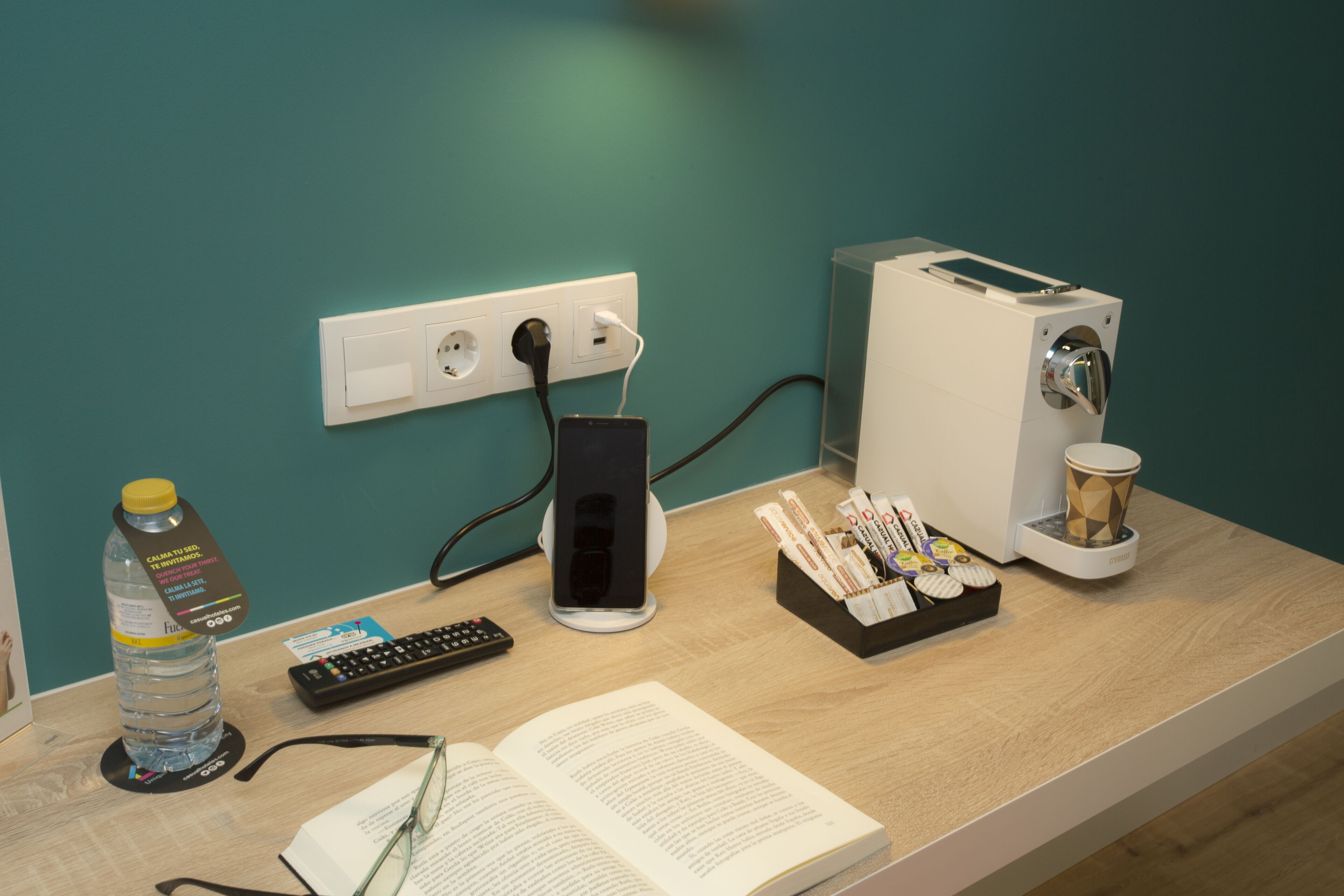 A desk with a laptop, smartphone, remote, book, glasses, water bottle, and a coffee machine.
