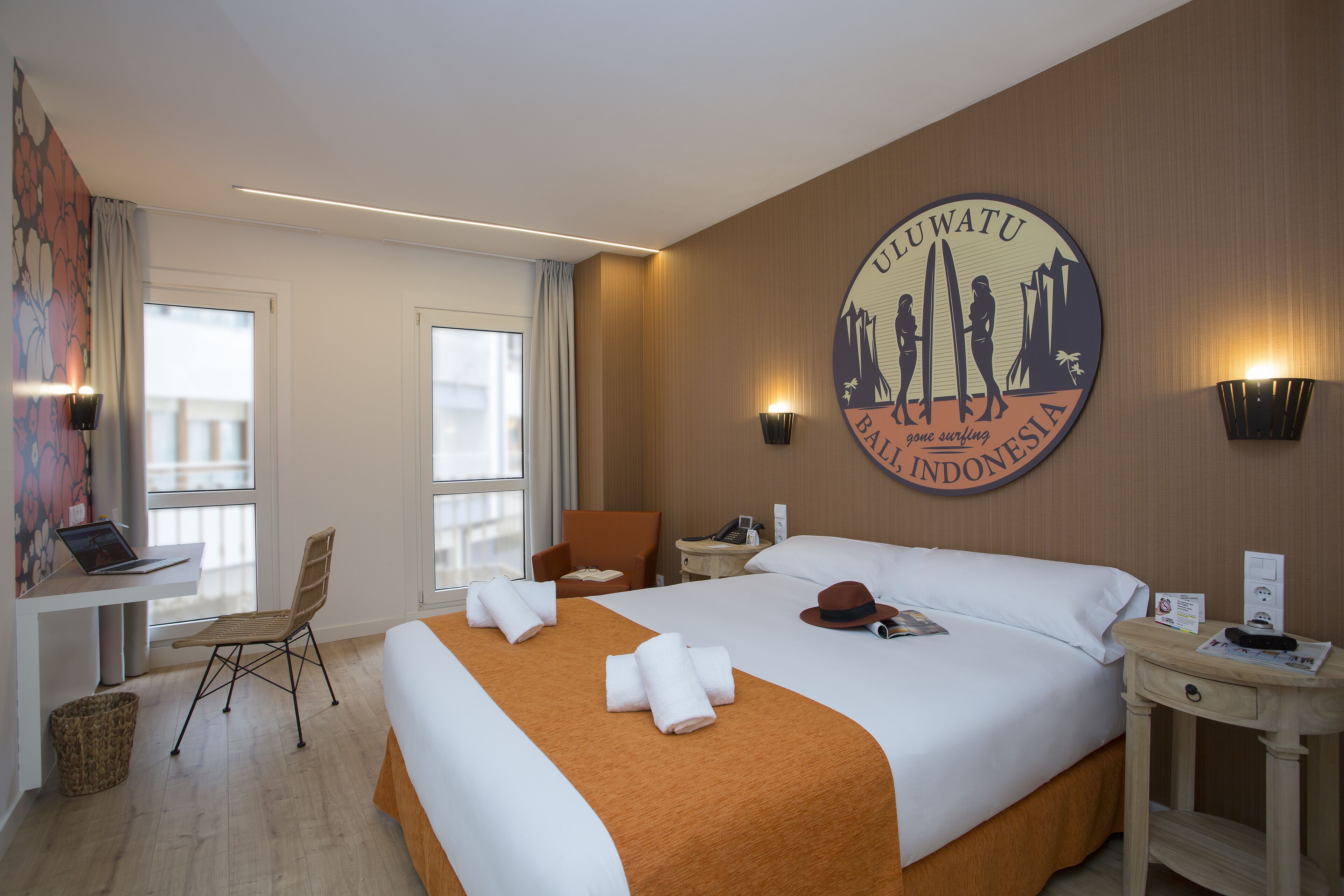 Una habitación de hotel con una cama grande, un escritorio con una computadora portátil, una silla, una mesita de noche y una pared con un logotipo de temática surf.
