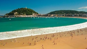Ein Strand mit einer großen Menschenmenge, klarem türkisblauem Wasser und einer Hügelkette mit Gebäuden im Hintergrund.