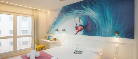 A hotel room with a large bed, a painting of a surfer on a wave, a small side table, and a window with curtains.