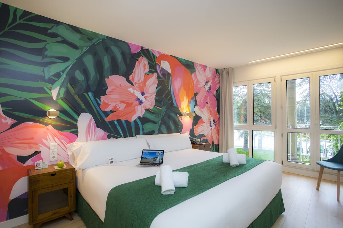 Une chambre d’hôtel avec un grand décor mural floral, un lit recouvert d’une couverture verte, un ordinateur portable posé sur le lit et une chaise près de la fenêtre.