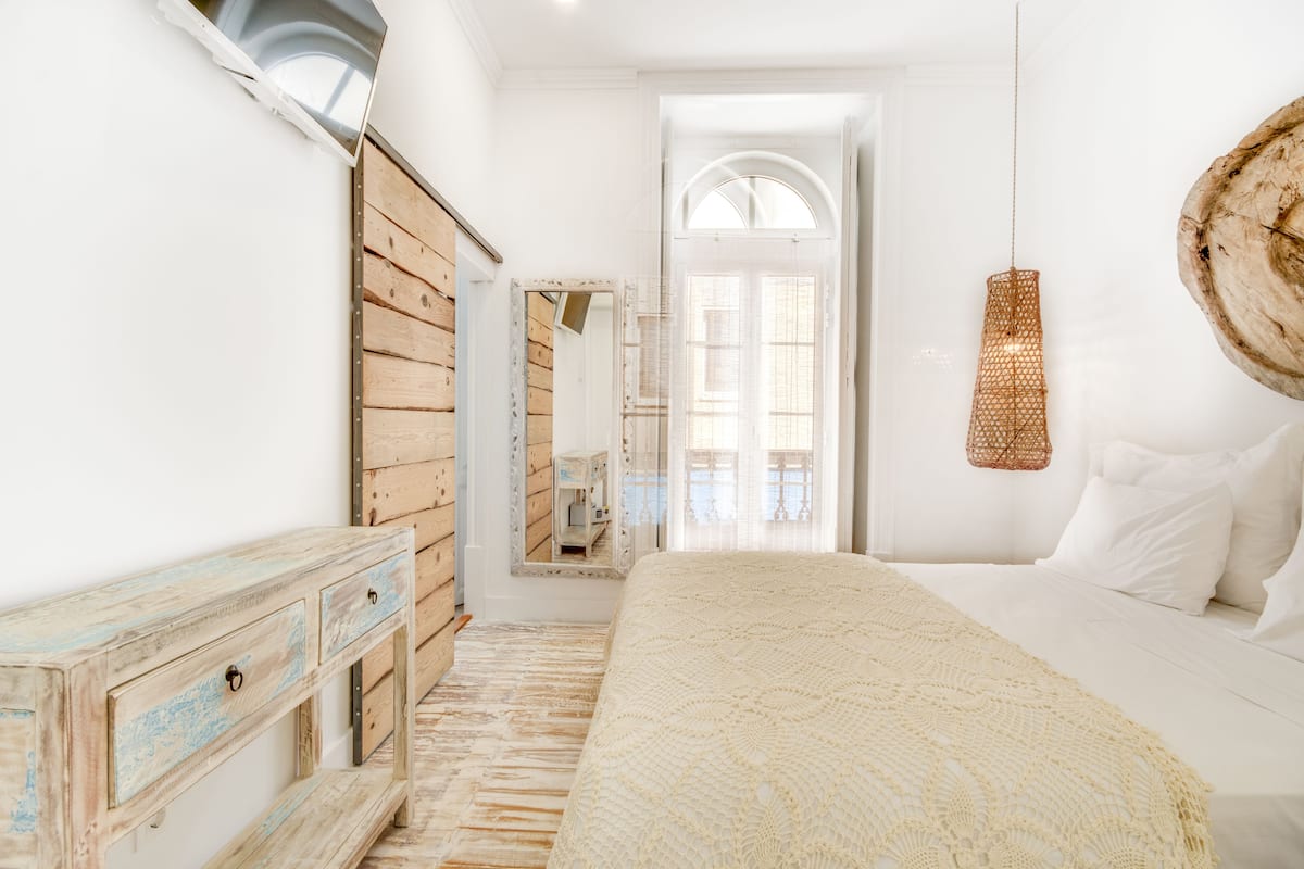 A bedroom with a wooden dresser, a large bed with a white blanket, a hanging light fixture, and a mirror on the wall.