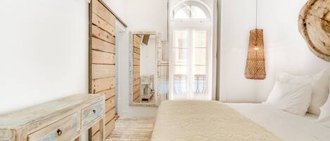 A bedroom with a wooden dresser, a large bed with a white blanket, a hanging light fixture, and a mirror on the wall.