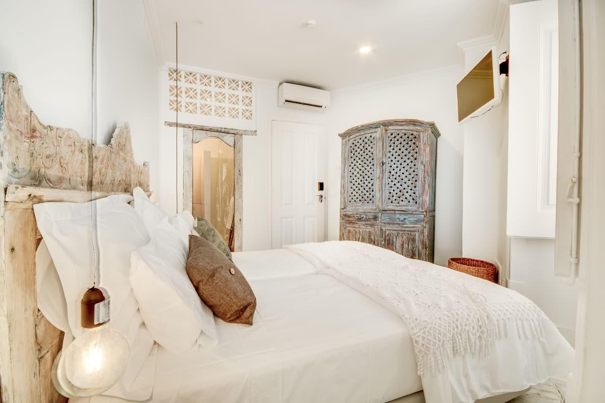 A bedroom with a bed, white bedding, a wooden headboard, a hanging light fixture, and a vintage wardrobe.