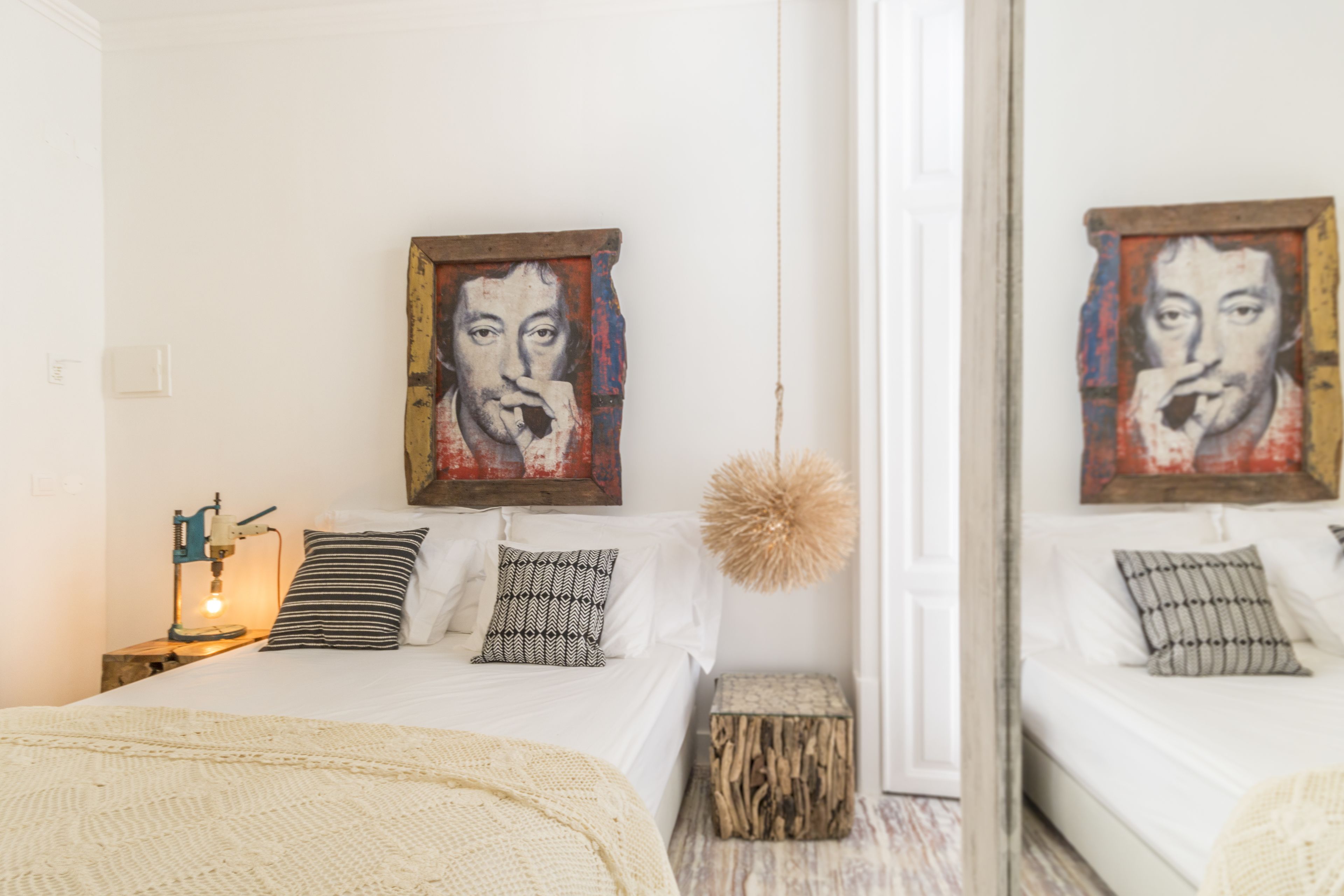 A bedroom with a bed, pillows, a bedside lamp, a small wooden stool, and a mirror.