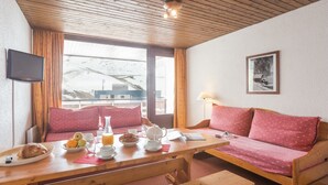 Una habitación acogedora con techo de madera, un sofá con cojines rojos, una mesa con alimentos para el desayuno y una ventana con vistas a un paisaje nevado.
