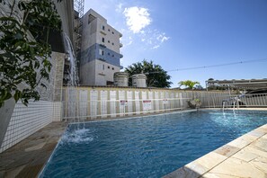 Piscina com parede revestida, fonte de água e um edifício com vários andares ao fundo.