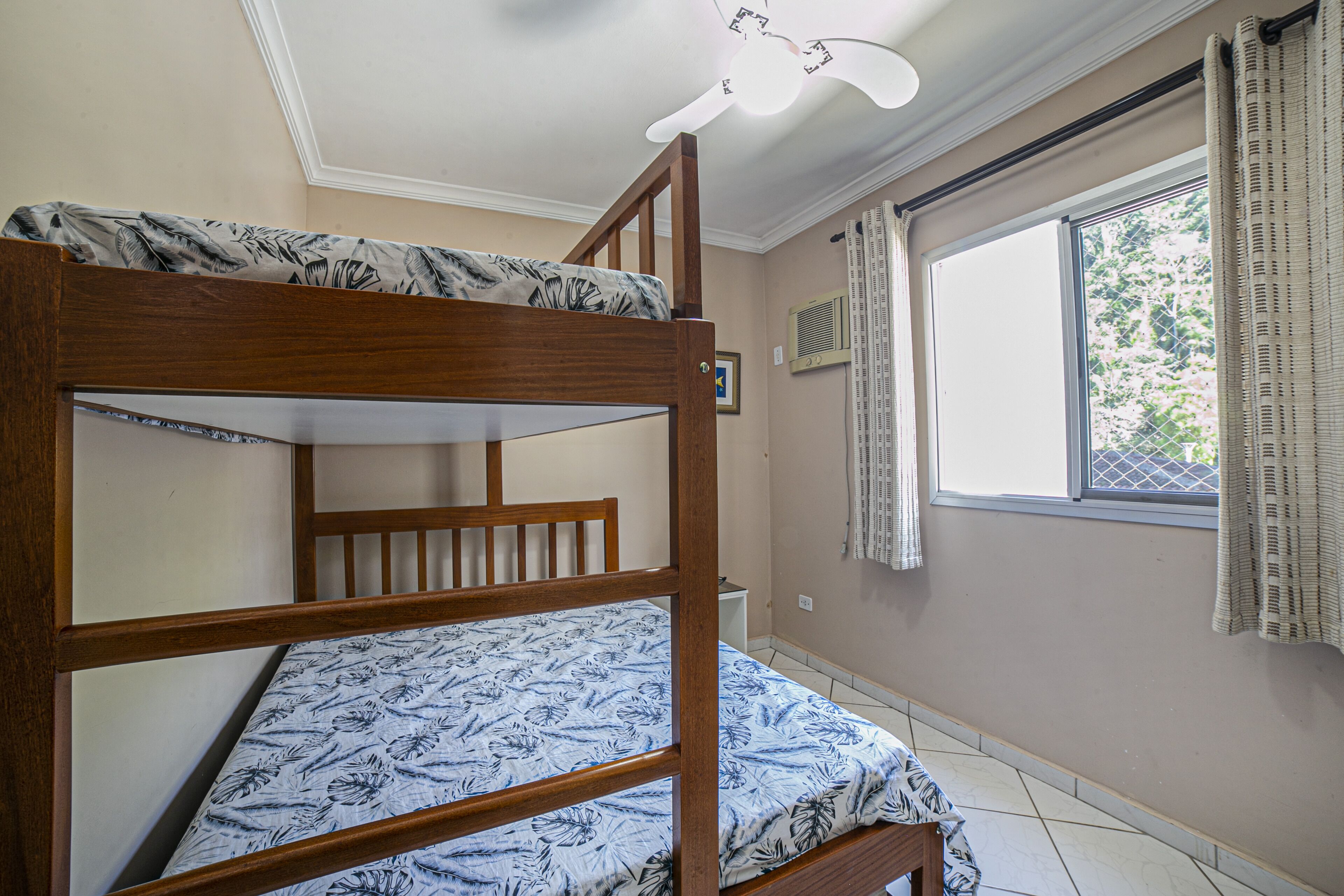 Uma cama beliche com edredom estampado, uma janela com cortinas e um lustre no teto.