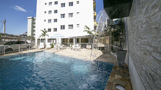A swimming pool with a water feature, surrounded by a patio area with outdoor seating and a building in the background. - Apto 22E ar condicionado e ventilador, 2 quartos, churrasqueira, elevador e piscina (Ubatuba)