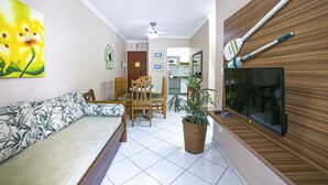 A living room with a sofa, a dining table with chairs, a TV mounted on the wall, and a potted plant. - Apto 22E ar condicionado e ventilador, 2 quartos, churrasqueira, elevador e piscina (Ubatuba)