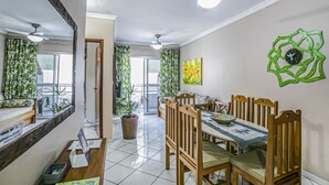 A dining area with wooden chairs and a table, a ceiling fan, and a decorative wall piece. - Apto 22E ar condicionado e ventilador, 2 quartos, churrasqueira, elevador e piscina (Ubatuba)