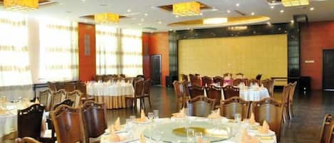Un salón de banquetes con mesas redondas dispuestas para comer, sillas de madera y un escenario al fondo.