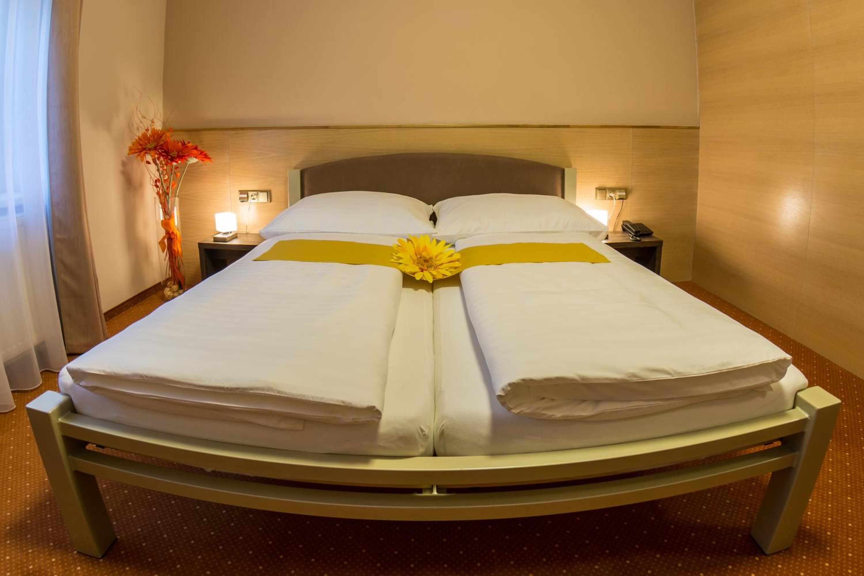 A double bed with white linens and a yellow sash, two bedside tables with lamps, and a vase with orange flowers on the left.