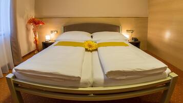 A double bed with white linens and a yellow sash, two bedside tables with lamps, and a vase with orange flowers on the left.