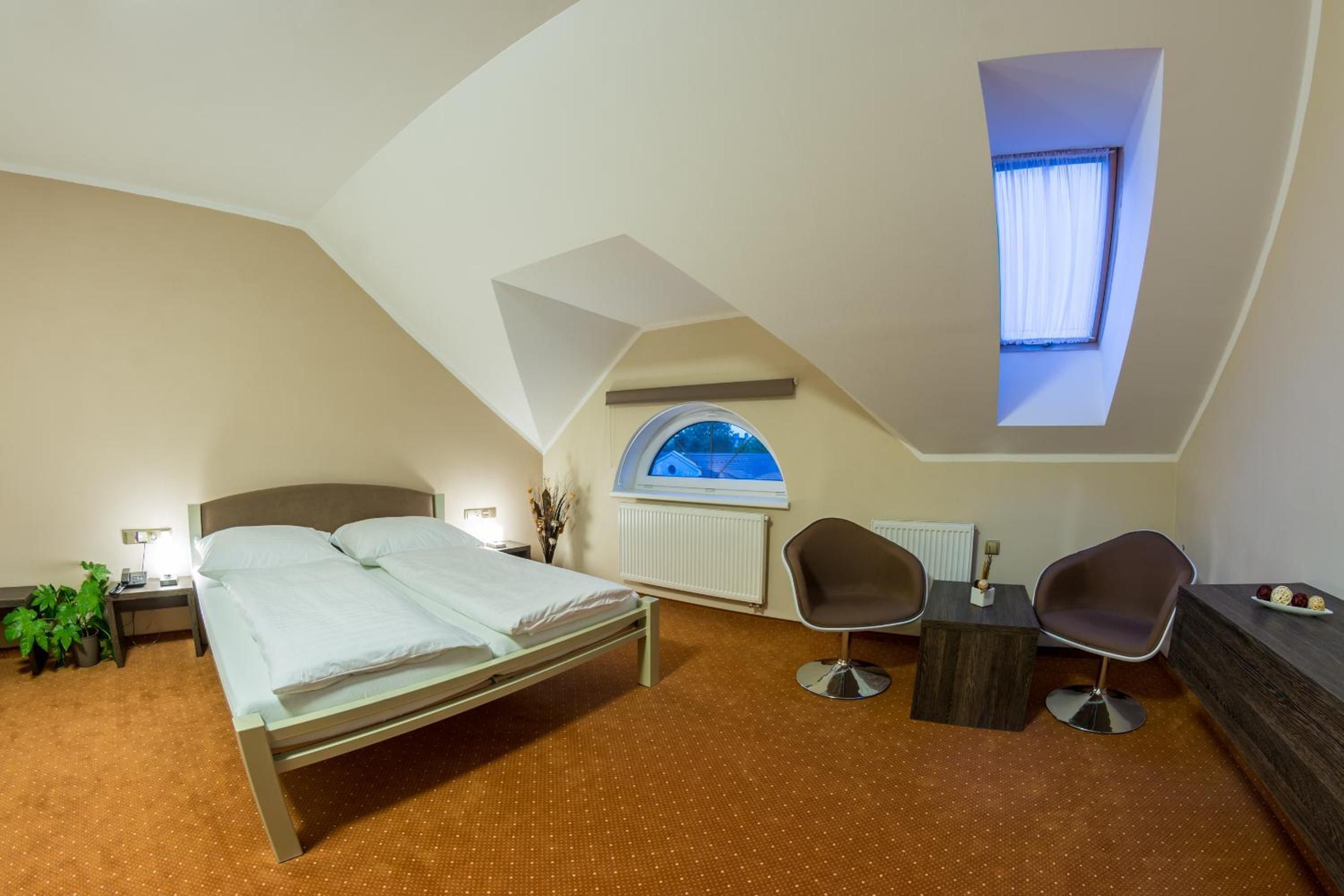 A bedroom with a bed, two chairs, a small table, a radiator, and a window with a view.