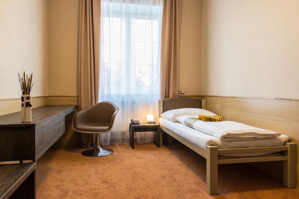 A hotel room with a single bed, a desk, a chair, a nightstand, and a window with curtains.