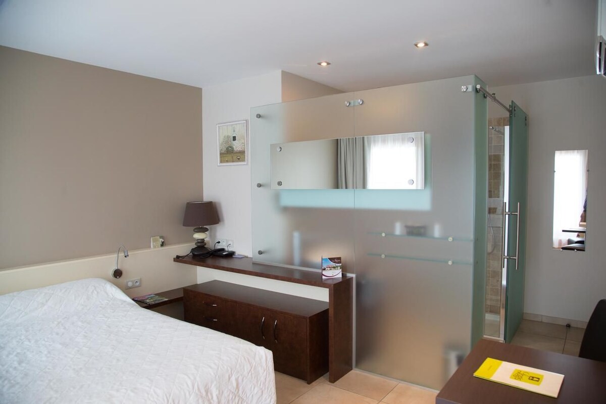 A hotel room with a bed, a desk with a lamp, a mirror, and a shower area.