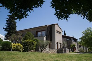 Una casa de dos pisos con fachada de piedra, un balcón en el nivel superior y un jardín cuidado con diversos arbustos y árboles.